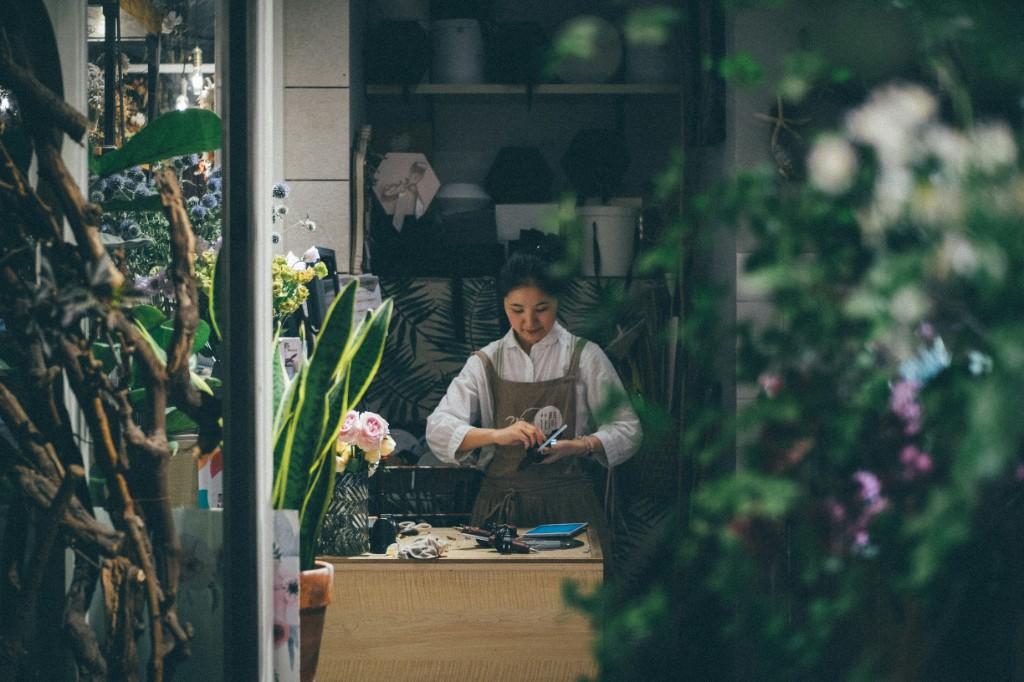 A shopkeeper at the counter, working on a phone among plants and flowers